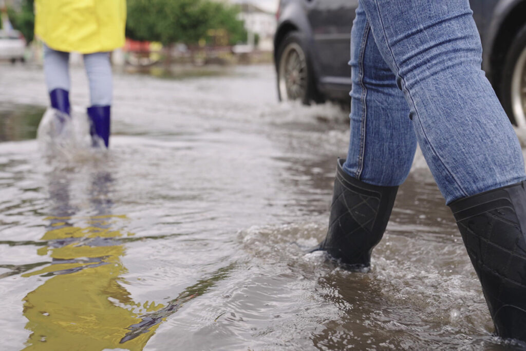 girls in raincoats and rubber boots walk along road flooded with torrential rains, their feet walk through puddles city, splashing water to the sides, the flood is on street, car is driving on water.